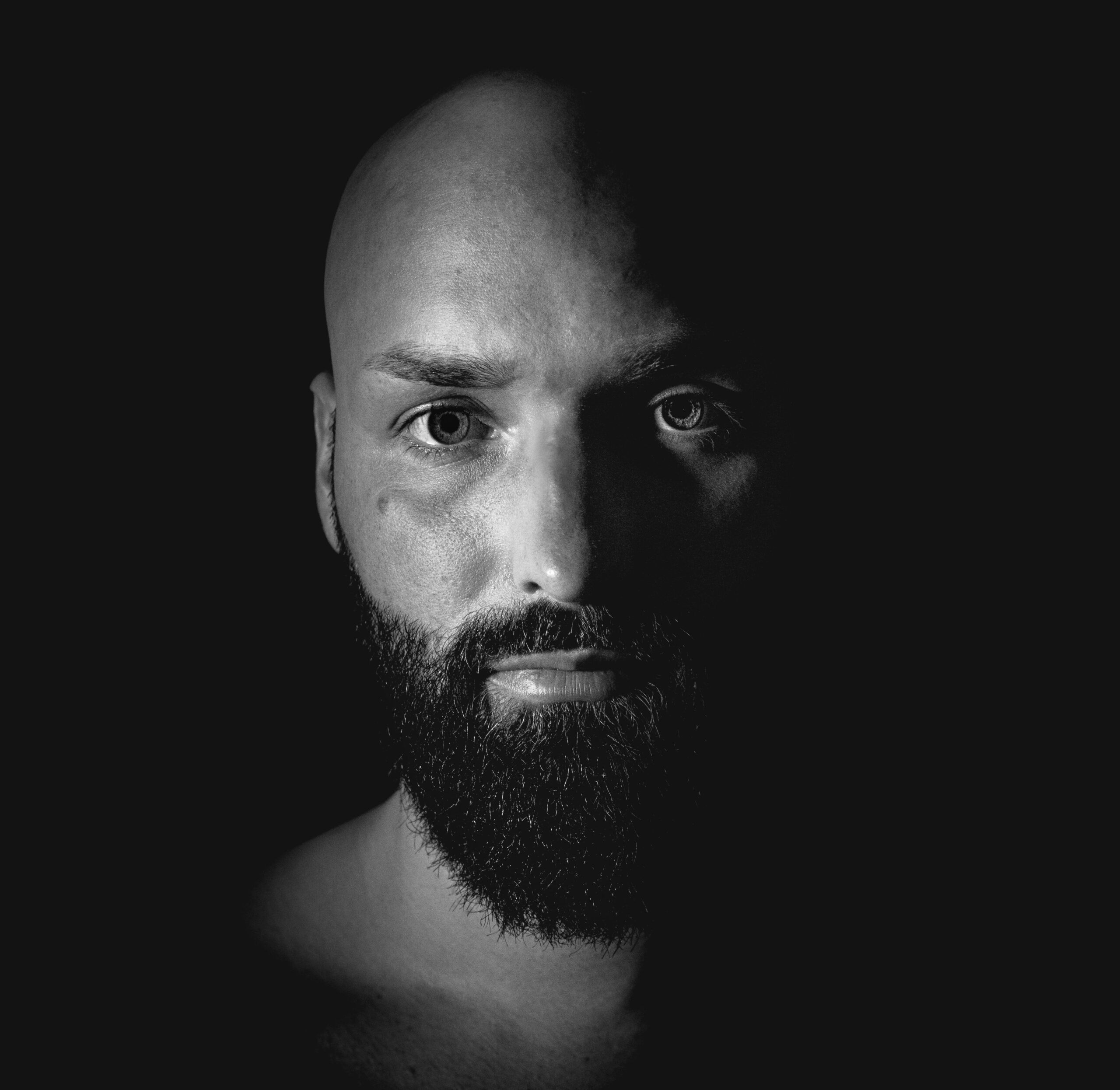testimonial-skip-01 Close-up black and white portrait of a bald man with a beard, highlighting dramatic shading and facial features.